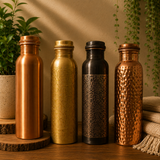 Four decorative metal water bottles in copper, gold, black, and hammered copper finishes, arranged on a wooden surface with plants and folded fabric in the background.