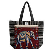A tote bag featuring a large embroidered elephant motif on a deep red front panel, flanked by black and white geometric patterned side panels, with black shoulder straps.