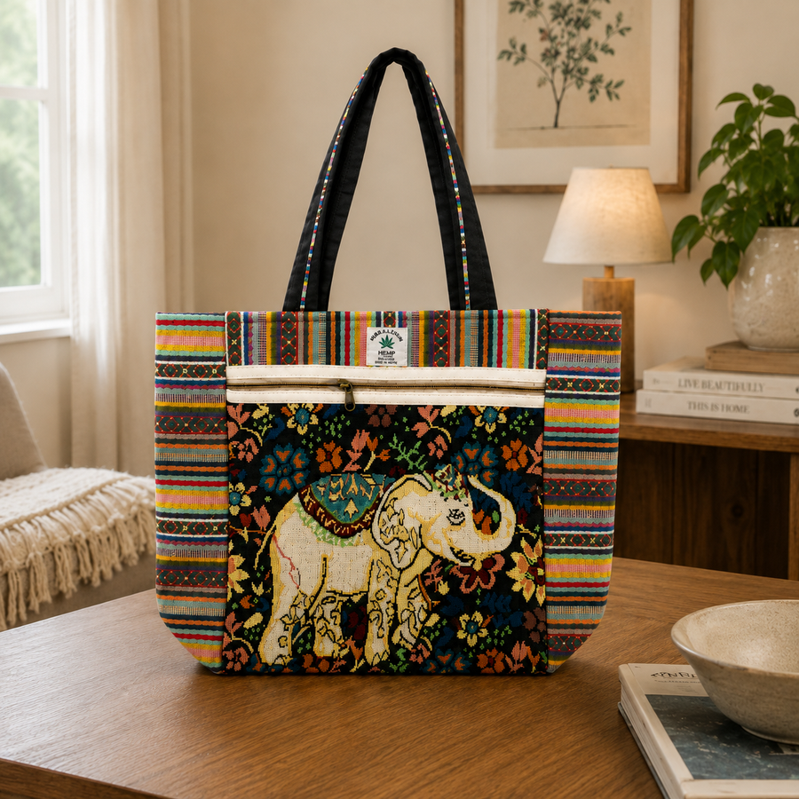 A woven tote bag with a colorful striped border, a black handle, and a central panel featuring a cream elephant on a dark floral background. The bag is displayed on a wooden table in a cozy, sunlit room with a lamp, books, and a plant in the background.