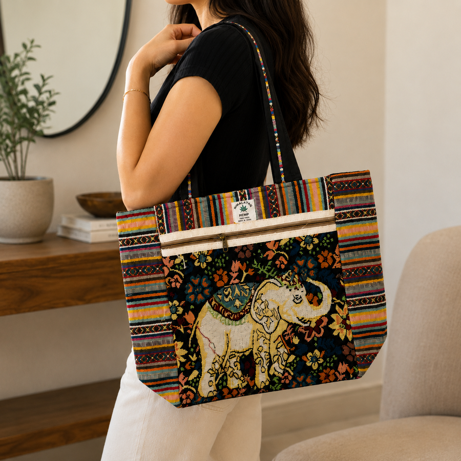 A woman wearing a black top and white pants carries a large tote bag with a colorful striped border and a central elephant illustration on a dark floral background.