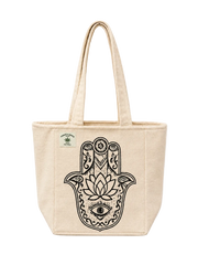 A natural beige canvas tote bag with a large black Hamsa hand graphic featuring a lotus flower and eye design on the front. A small green brand label is visible on the upper left side.