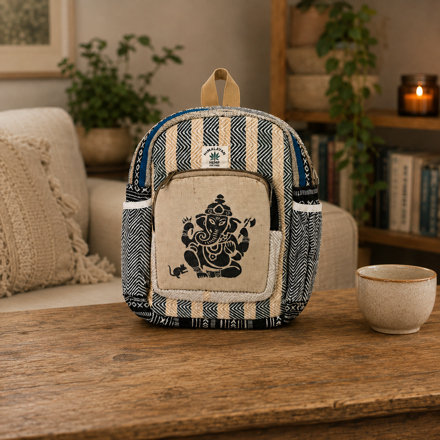 A small backpack made from cream and navy patterned fabric sits on a wooden table in a cozy living room setting. The front pocket features a black printed silhouette of Ganesha, and a small white label with a green leaf logo is stitched near the top.