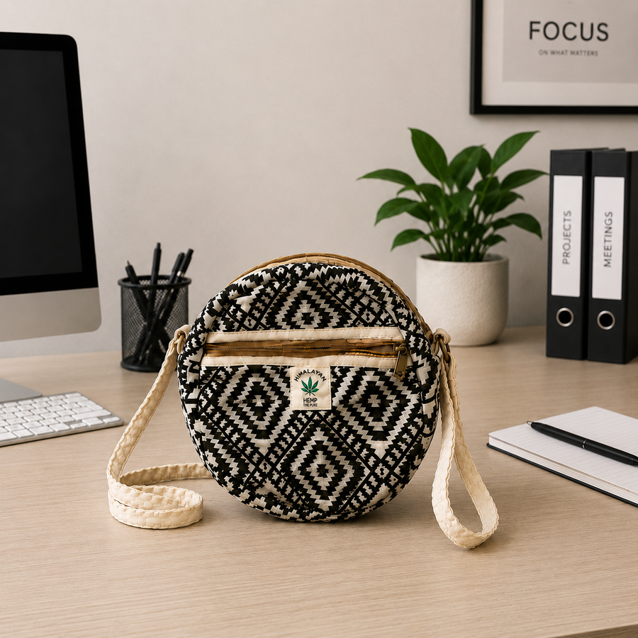 A round woven crossbody bag with a black and cream geometric pattern, resting on a light wood desk in a styled office environment with a computer monitor, plant, and binders in the background.