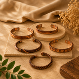 Five copper-toned cuff bracelets with various geometric and textured patterns, arranged on a beige stone surface with a draped beige fabric and dried flowers in the background.