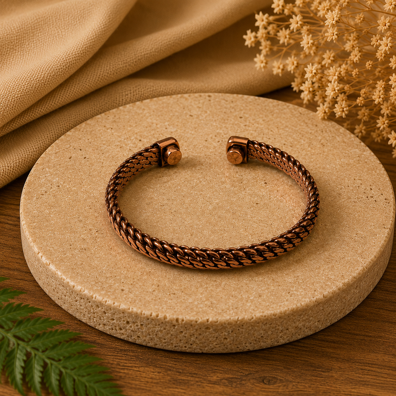 A twisted rope-style cuff bracelet in a warm copper or rose-gold tone, displayed on a round beige stone slab with draped fabric and dried flowers in the background.